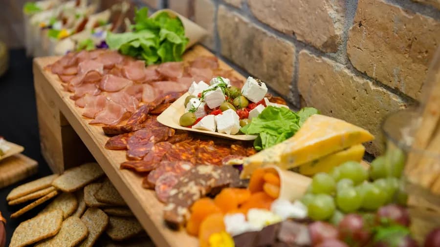 A long wooden grazing stand of cured meats, cheeses, crackers and grapes against a brick wall