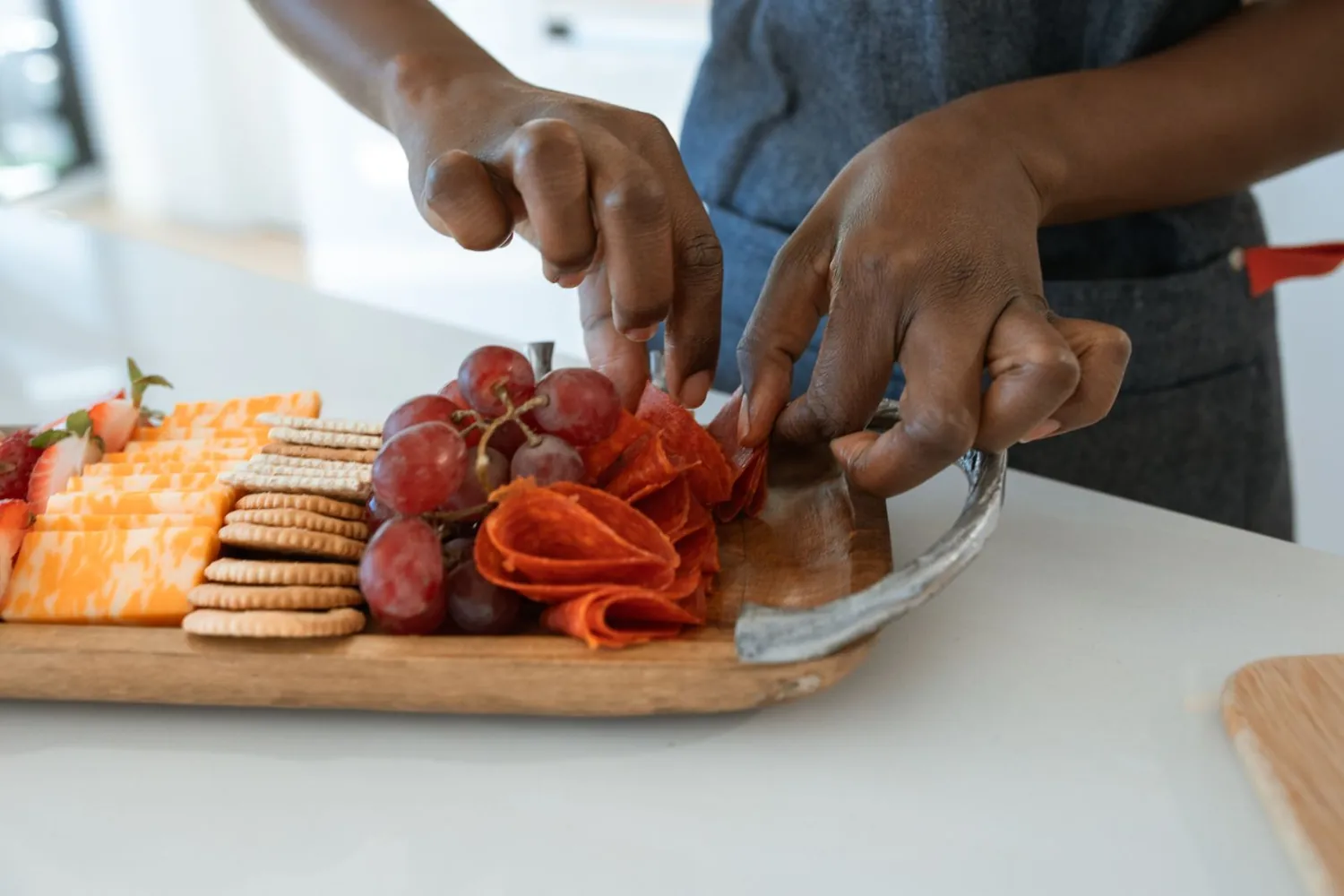 Hands arranging folded salami, grapes, crackers and sliced cheese on a long wooden board