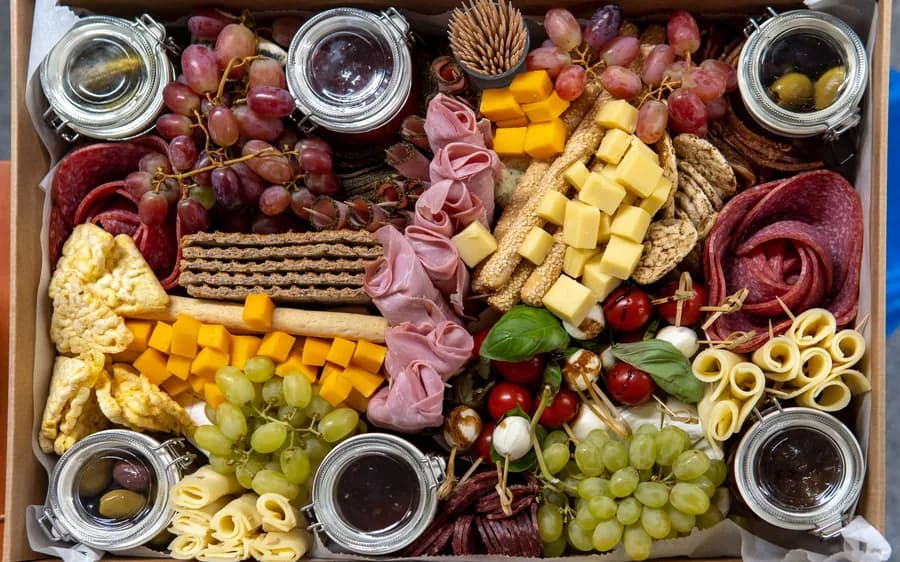 A grazing box packed with cured meats, cheeses, grapes, crackers and small jars of olives and jam