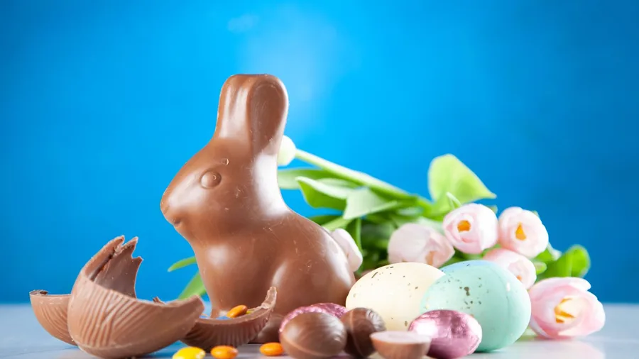 A chocolate Easter bunny beside a cracked chocolate egg, pastel eggs and pink tulips