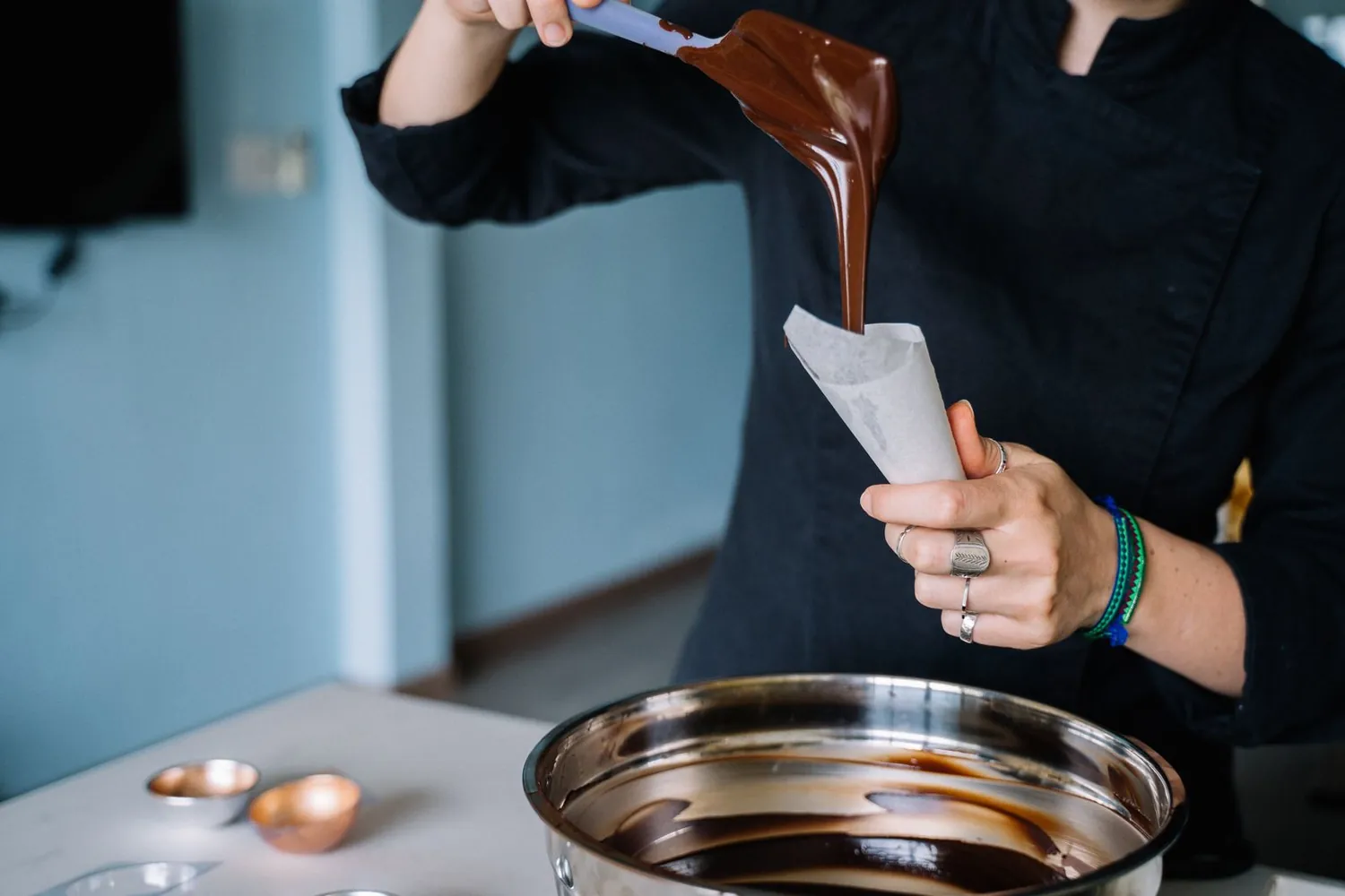 A chocolatier in a black jacket pouring melted chocolate into a piping bag over a steel bowl